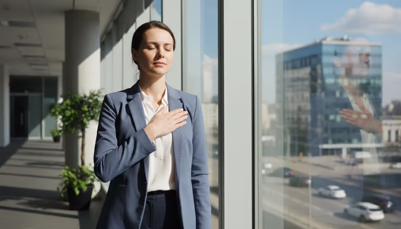 Woman standing by an office window with eyes closed and hand on her heart, taking a brief mindfulness pause during a workday, cityscape visible through the window
