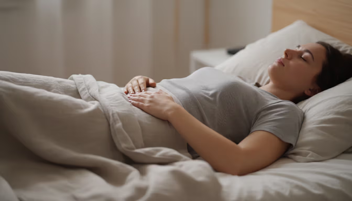 Person lying in bed practicing diaphragmatic breathing with one hand on chest and one hand on abdomen in a softly lit room with a pillow under knees