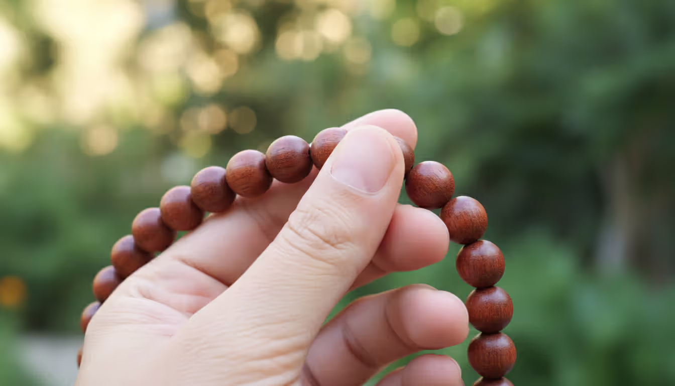 Close-up of a hand holding and counting wooden mala beads with 108 beads against a blurred green garden background in warm natural light
